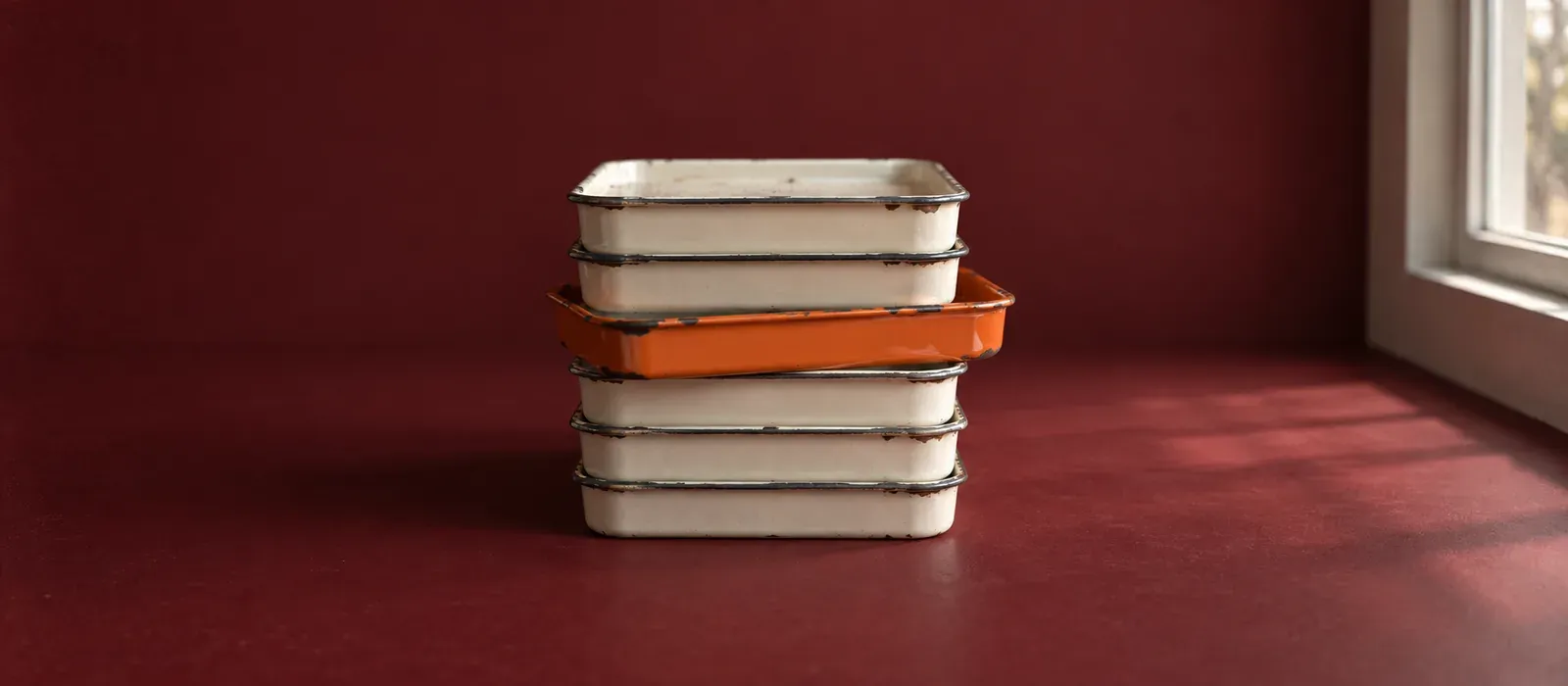 A stack of six enamelled steel trays on an oxblood ground, five bone-white with chipped rims, the third from the top drawn out of the column and glazed orange.