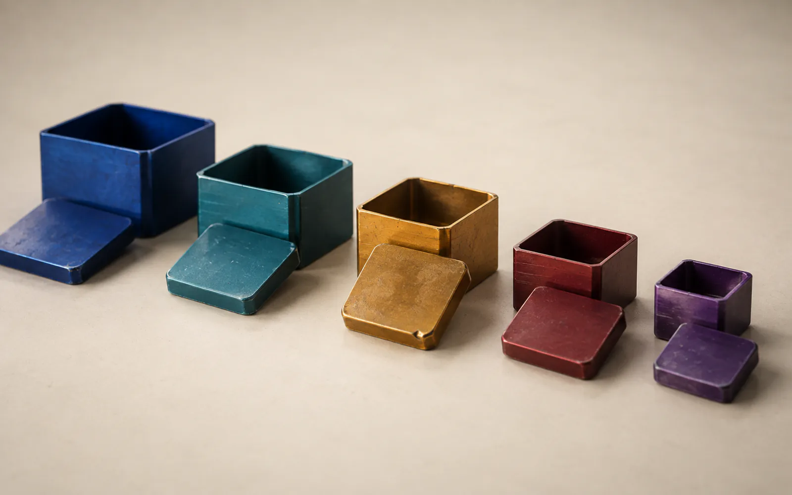 Five lidded ceramic boxes in descending sizes — blue, teal, ochre, red and violet — set in a row on a cream ground.